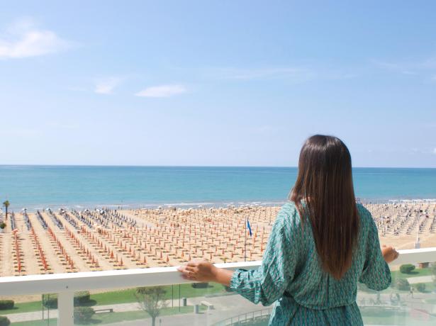 Frau blickt vom Balkon aufs Meer, Strand mit Sonnenschirmen.